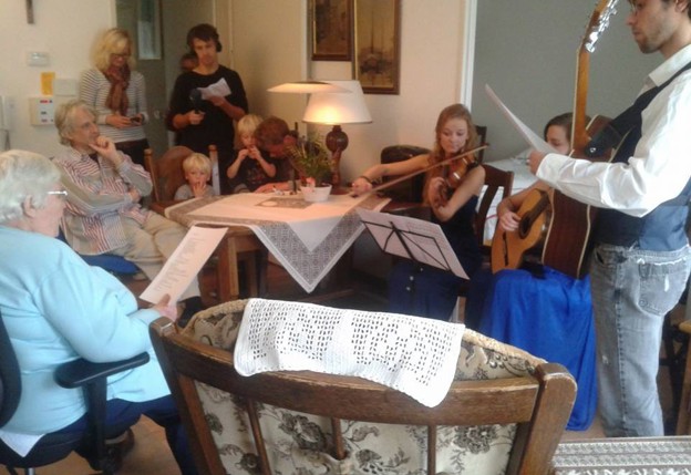 Mensen van verschillende leeftijden luisteren in een huiskamer naar muzikanten die viool en gitaar spelen.