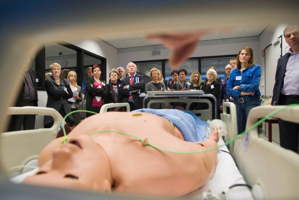 Een groep mensen observeert aandachtig een medische demonstratie met een levensechte oefenpop op een ziekenhuisbed. De pop is aangesloten op slangen en kabels, en de demonstratie vindt plaats in een professionele opleidingsruimte.