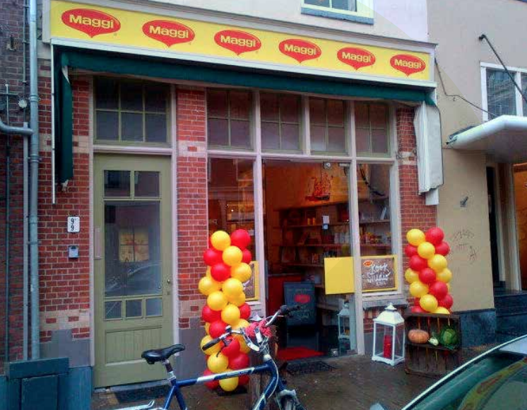 Een winkelpui met een groen-wit gestreepte luifel waarop meerdere keren het woord "Maggi" staat. Voor de pui staan twee trossen ballonnen in rood en geel, en een blauwe fiets.