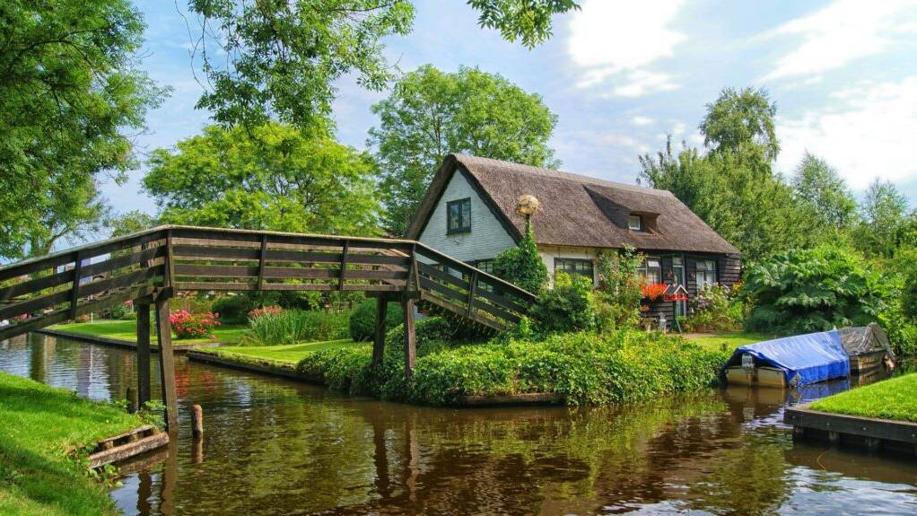 en pittoresk huis met een rieten dak, gelegen aan een rustige waterweg in Giethoorn, omringd door groen en verbonden door een houten bruggetje.