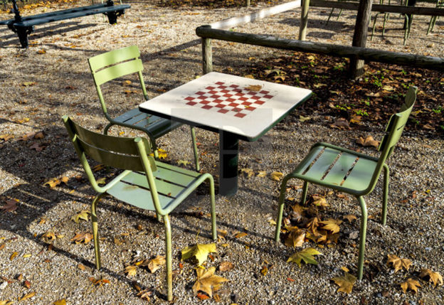 Een vierkante schaaktafel met een rood-wit geblokt schaakbord staat buiten op een terras met vier groene metalen stoelen eromheen. De tafel is leeg en er zijn geen schaakstukken te zien. Op de achtergrond is een hekwerk van houten palen zichtbaar, evenals de schaduw van bomen op de grond met gevallen bladeren.