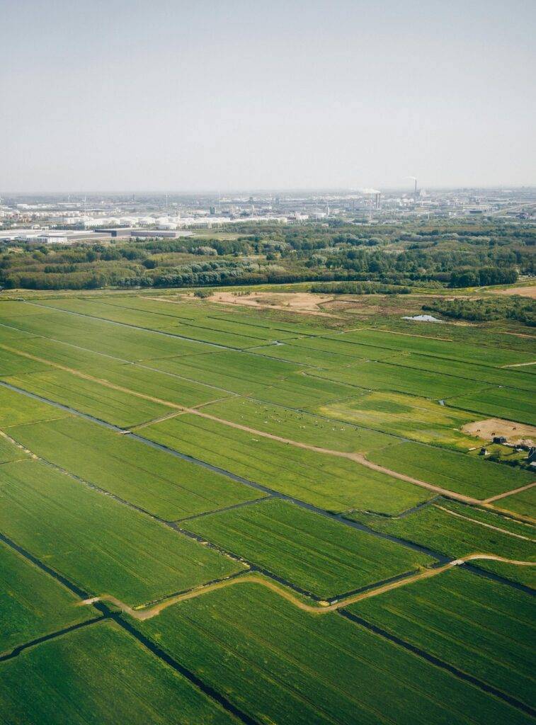 Een luchtfoto van een uitgestrekt landbouwgebied met groene velden, doorsneden door rechte sloten. Op de achtergrond is een stad te zien met fabrieken en bebouwing. De lucht is helder en de zon schijnt, wat zorgt voor een rustige en weidse sfeer.