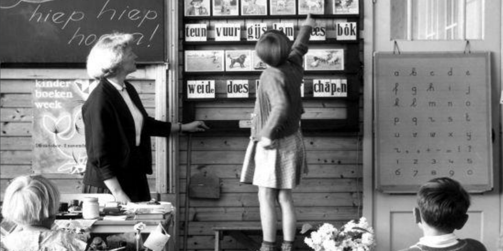 Foto uit jaren vijftig van een onderwijzeres die voor de klas staat. Ze geeft taalles.