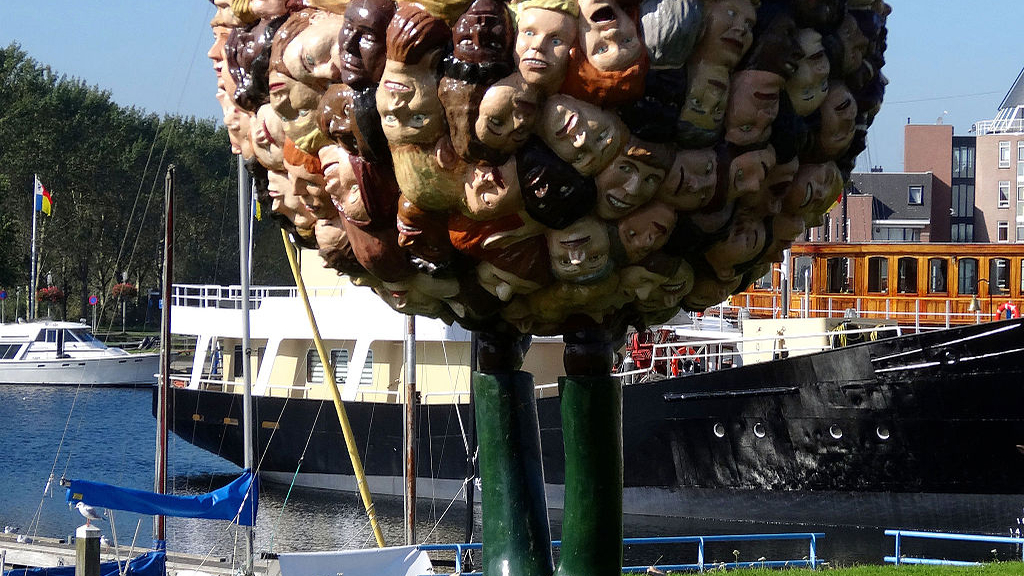 Beeldhouwwerk van Paul de Reus in Almere Haven, bestaande uit een boomvorm met tientallen kleurrijke hoofden, met op de achtergrond een haven met boten en gebouwen.