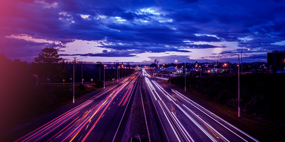 Een lange weg met lichtsporen van auto's onder een donkere, bewolkte avondhemel.