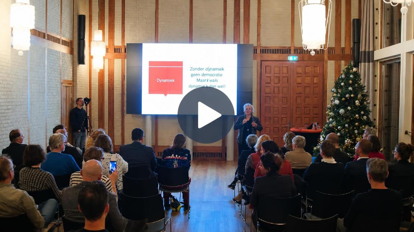 Filmstill van de lezing met de spreker voor een scherm in een zaal met mensen waar ook een kerstboom staat.