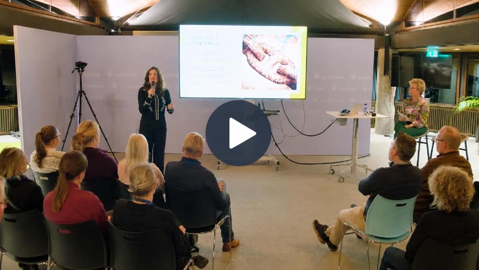 Filmstill van de lezing met de spreker naast een scherm voor een zaal met mensen.