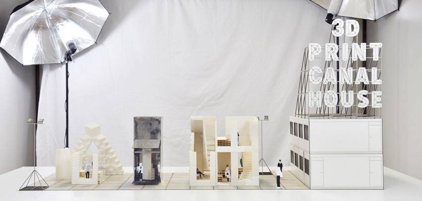 Een architectonisch model van een 3D-geprint grachtenpand, tentoongesteld in een studio-opstelling met professionele belichting. Het model toont verschillende bouwfasen en constructietechnieken. Rechts staat een groot bord met de tekst "3D PRINT CANAL HOUSE." Miniatuurfiguren verkennen de ruimtes, wat de schaal en functionaliteit van het ontwerp benadrukt.