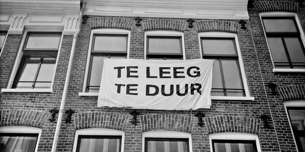 Gevel van oud pand waaraan spandoek hangt met tekst 'Te leeg. Te duur'