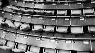 Een zwart-wit foto van bovenaf toont een lege vergaderzaal met rijen identieke stoelen en tafels