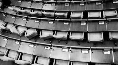 Een zwart-wit foto van bovenaf toont een lege vergaderzaal met rijen identieke stoelen en tafels